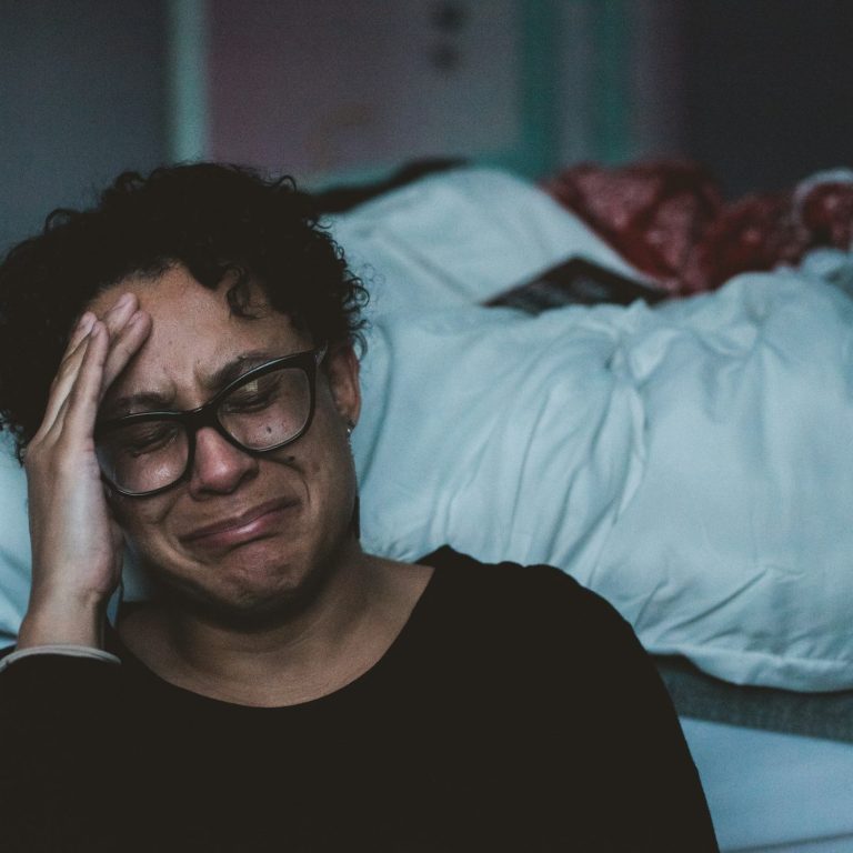 Mujer con gafas, visiblemente angustiada, sentada junto a una cama desordenada.